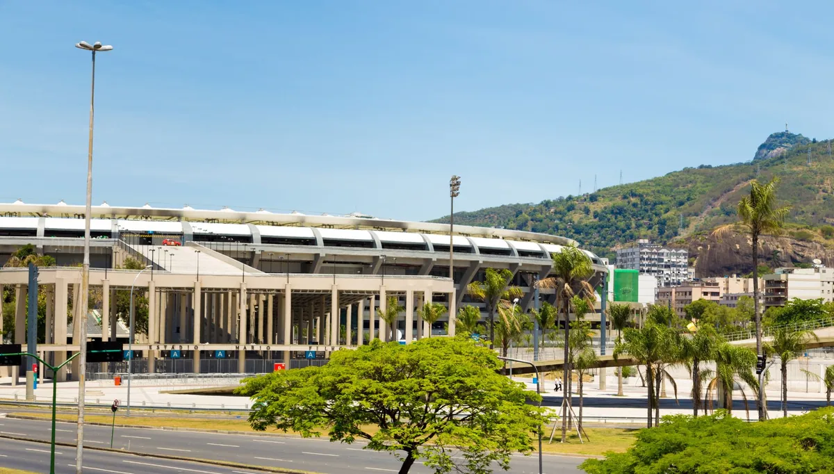 Fachada de estádio de futebol em dia claro, com morro ao fundo